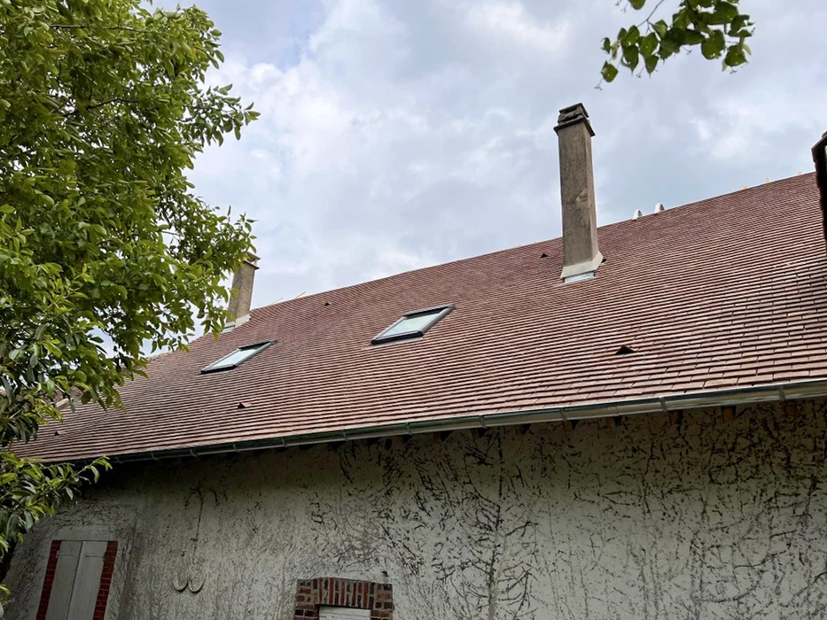 Extérieur d'une maison traditionnelle avec mur crépi blanc fissuré, toiture en tuiles terre cuite avec cheminées en pierre et lucarnes