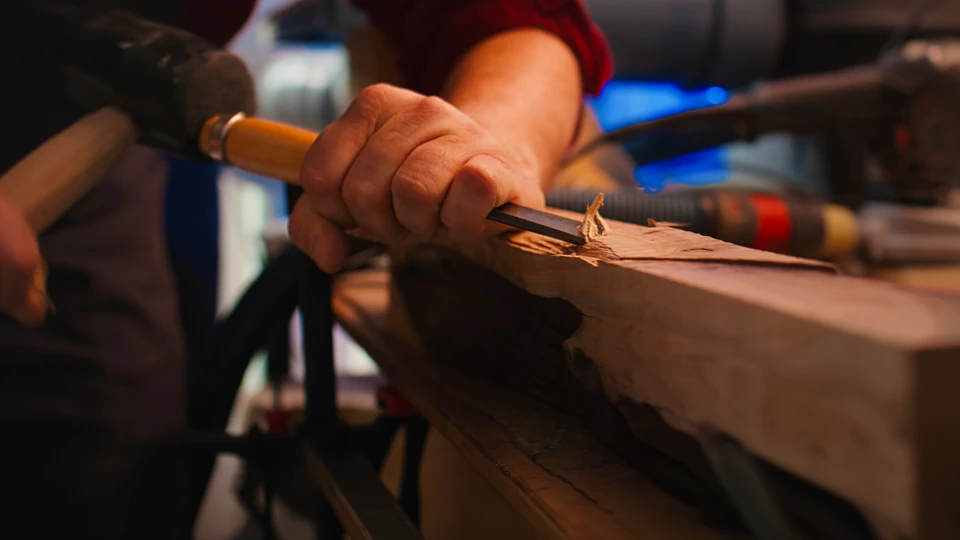 Atelier de menuiserie intérieur où un artisan en pull beige utilise une mortaiseuse à bédane pour percer des mortaises dans une pièce de bois massif